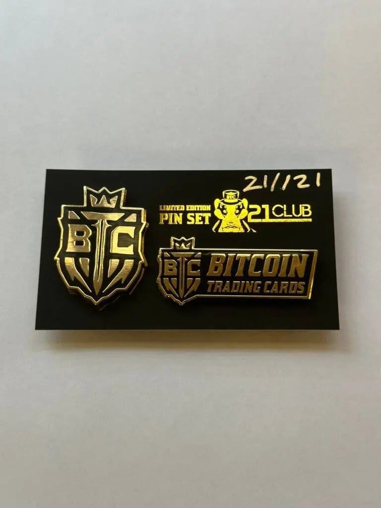 Close-up photograph of the '21 Club' Limited Edition Pin Set on a black backing card, numbered '21 / 21' in gold marker. Includes a gold and black shield pin and a rectangular 'Bitcoin Trading Cards' logo pin.