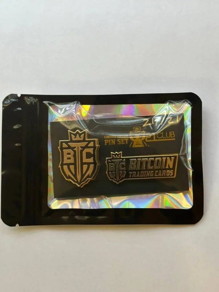 Photograph of the '21 Club' Limited Edition Pin Set (21/21) sealed inside black packaging with a clear window and holographic background, highlighting the gold pins and hand-numbered markings.