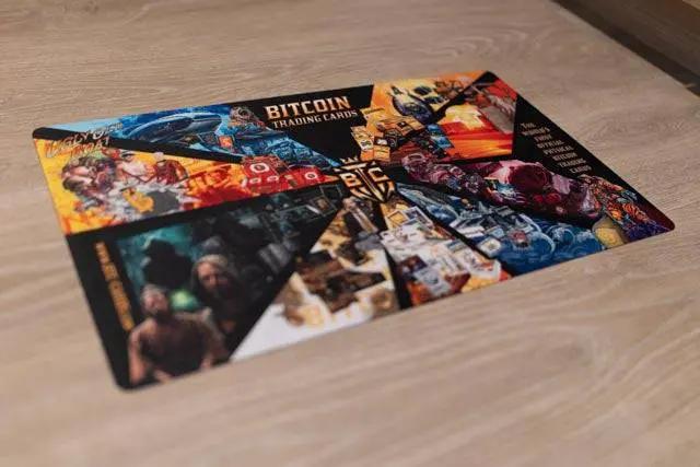 Photo of the official 'Mashup' breaker mat for Bitcoin Trading Cards lying on a wooden table, featuring a vibrant collage of series artwork including the Whale, Crab, and Ugly Old Goat characters.