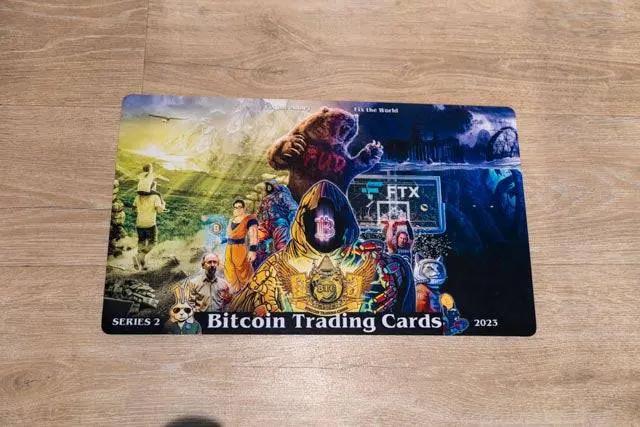 Top-down photograph of the physical Series 2 breaker mat lying on a wooden floor, displaying the full 'FUD Busters' artwork including the bear, tombstone, and 2023 date.