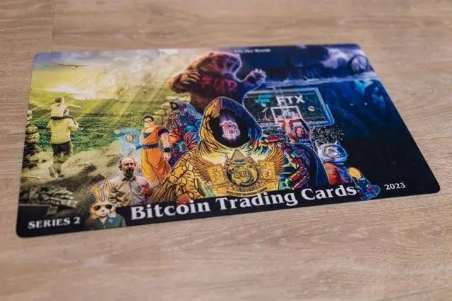 Photograph of the physical Series 2 breaker mat for Bitcoin Trading Cards lying on a wooden floor, clearly showing the 'FUD' bear, FTX tombstone, and 2023 date in a real-world setting.