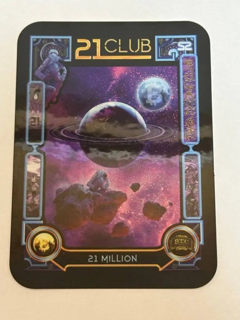 Close-up photograph of the limited edition '21 Club' sticker titled '21 Million', featuring a space scene with an astronaut sitting on an asteroid, a floating astronaut, a planet with rings, and a moon embossed with the Bitcoin logo.
